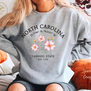 Op de afbeelding: Een lichtgrijs sweatshirt met de tekst "NORTH CAROLINA" en co&ouml;rdinaten, met roze bloemenillustraties. Daaronder staat "TARHEEL STATE EST. 1789". Een comfortabele, casual stijl.