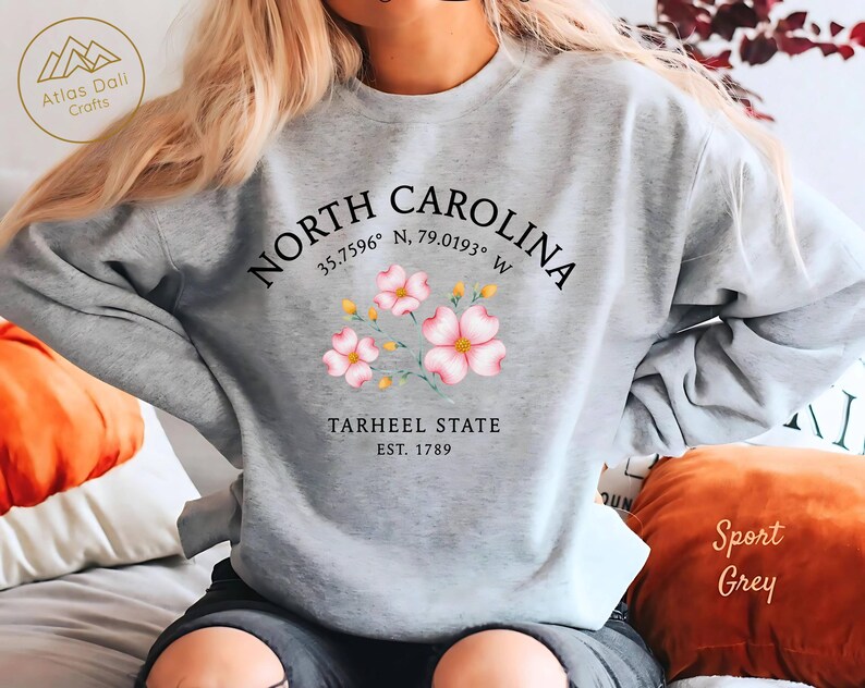 Op de afbeelding: Een lichtgrijs sweatshirt met de tekst "NORTH CAROLINA" en co&ouml;rdinaten, met roze bloemenillustraties. Daaronder staat "TARHEEL STATE EST. 1789". Een comfortabele, casual stijl.