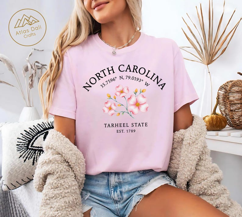 Może przedstawiać: Jasnor&oacute;żowa koszulka z napisem "North Carolina", wsp&oacute;łrzędnymi, "Tarheel State" i "Est. 1789". Koszulka ma kwiatowy wz&oacute;r w kolorze r&oacute;żowym i białym. Logo marki "Atlas Dali Crafts" znajduje się w lewym g&oacute;rnym rogu.