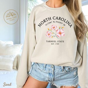Op de afbeelding: Zandkleurige sweatshirt met de tekst "North Carolina" en co&ouml;rdinaten, een bloemenafbeelding en "Tarheel State Est. 1789". De sweatshirt heeft een ronde hals.
