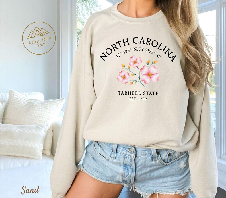 Op de afbeelding: Zandkleurige sweatshirt met de tekst "North Carolina" en co&ouml;rdinaten, een bloemenafbeelding en "Tarheel State Est. 1789". De sweatshirt heeft een ronde hals.