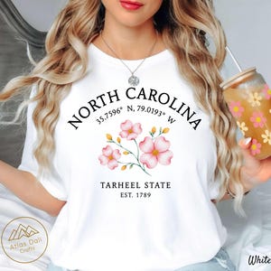 Może przedstawiać: Biała koszulka z napisem "NORTH CAROLINA" i kwiatowym wzorem z r&oacute;żowymi kwiatami. Na koszulce wydrukowano r&oacute;wnież słowa "TARHEEL STATE EST. 1789". W dłoni trzymany jest przezroczysty szklany kubek z kwiatowym wzorem.