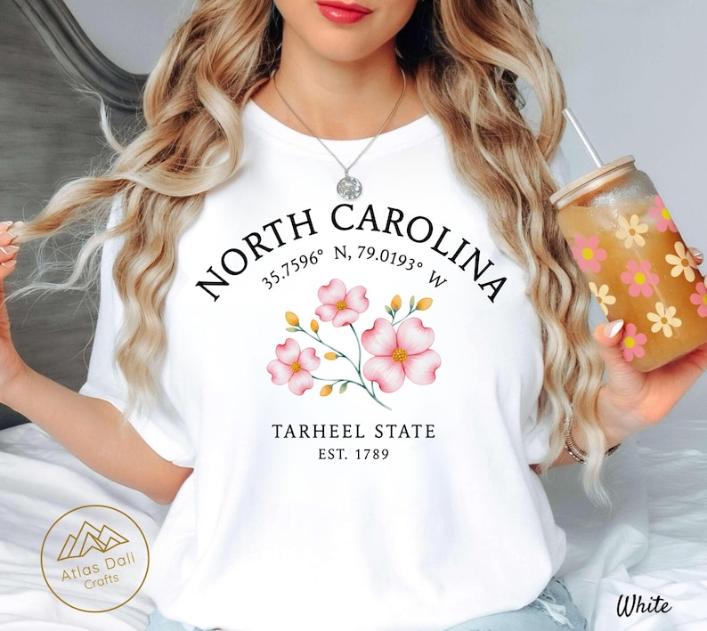 Może przedstawiać: Biała koszulka z napisem "NORTH CAROLINA" i kwiatowym wzorem z r&oacute;żowymi kwiatami. Na koszulce wydrukowano r&oacute;wnież słowa "TARHEEL STATE EST. 1789". W dłoni trzymany jest przezroczysty szklany kubek z kwiatowym wzorem.