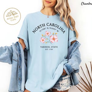 Może przedstawiać: Jasnoniebieska koszulka z napisem "NORTH CAROLINA" w okrągłym wzorze, wsp&oacute;łrzędnymi i "TARHEEL STATE EST. 1789". Koszulka posiada kwiatową grafikę w kolorze r&oacute;żowym i ż&oacute;łtym. Słowo "Chambray" jest wydrukowane w prawym g&oacute;rnym rogu.