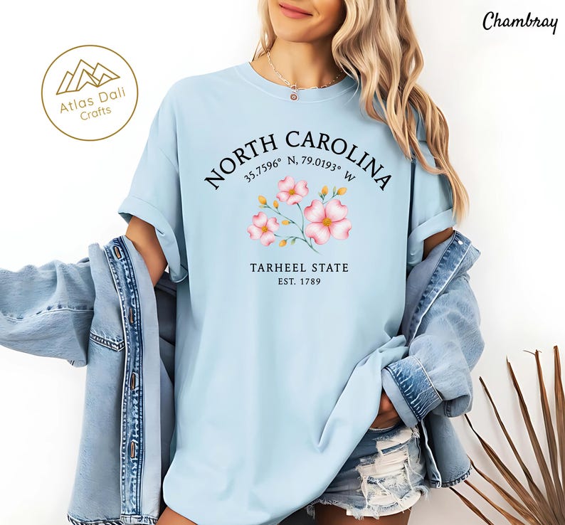 Może przedstawiać: Jasnoniebieska koszulka z napisem "NORTH CAROLINA" w okrągłym wzorze, wsp&oacute;łrzędnymi i "TARHEEL STATE EST. 1789". Koszulka posiada kwiatową grafikę w kolorze r&oacute;żowym i ż&oacute;łtym. Słowo "Chambray" jest wydrukowane w prawym g&oacute;rnym rogu.