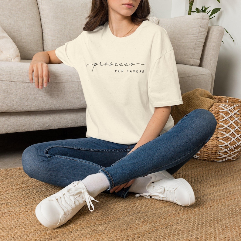Op de afbeelding: Een cr&egrave;mekleurig T-shirt met korte mouwen en een gespikkelde textuur. Op de voorkant staat de zwarte tekst "prosecco PER FAVORE" in een combinatie van cursieve en blokletters. Het T-shirt wordt gecombineerd met een donkerblauwe spijkerbroek en witte sneakers.