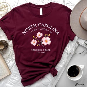 Może przedstawiać: Bordowa koszulka z napisem "NORTH CAROLINA", wsp&oacute;łrzędnymi i "TARHEEL STATE EST. 1789". Projekt zawiera element kwiatowy z r&oacute;żowymi kwiatami. Koszulka jest prezentowana na białej powierzchni, z filiżanką kawy i kapeluszem.