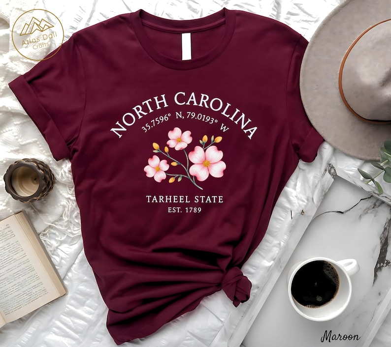Może przedstawiać: Bordowa koszulka z napisem "NORTH CAROLINA", wsp&oacute;łrzędnymi i "TARHEEL STATE EST. 1789". Projekt zawiera element kwiatowy z r&oacute;żowymi kwiatami. Koszulka jest prezentowana na białej powierzchni, z filiżanką kawy i kapeluszem.
