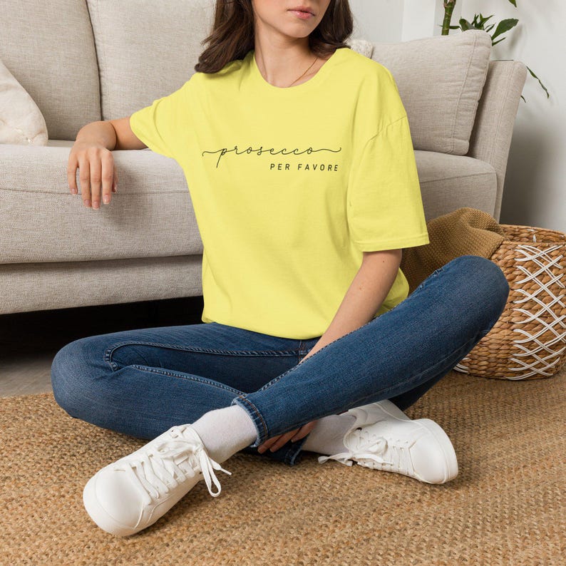Op de afbeelding: Een felgeel T-shirt met korte mouwen en de zwarte tekst "prosecco PER FAVORE" op de borst. Het shirt heeft een ronde hals en een relaxte pasvorm, gecombineerd met een donkerblauwe spijkerbroek en witte sneakers met veters.