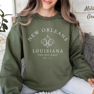 May include: Military green sweatshirt with the text "New Orleans Louisiana The Big Easy Est. 1718" in white. The sweatshirt features a fleur-de-lis design. The brand name "Atlas Daffi Crafts" is visible on the left side.