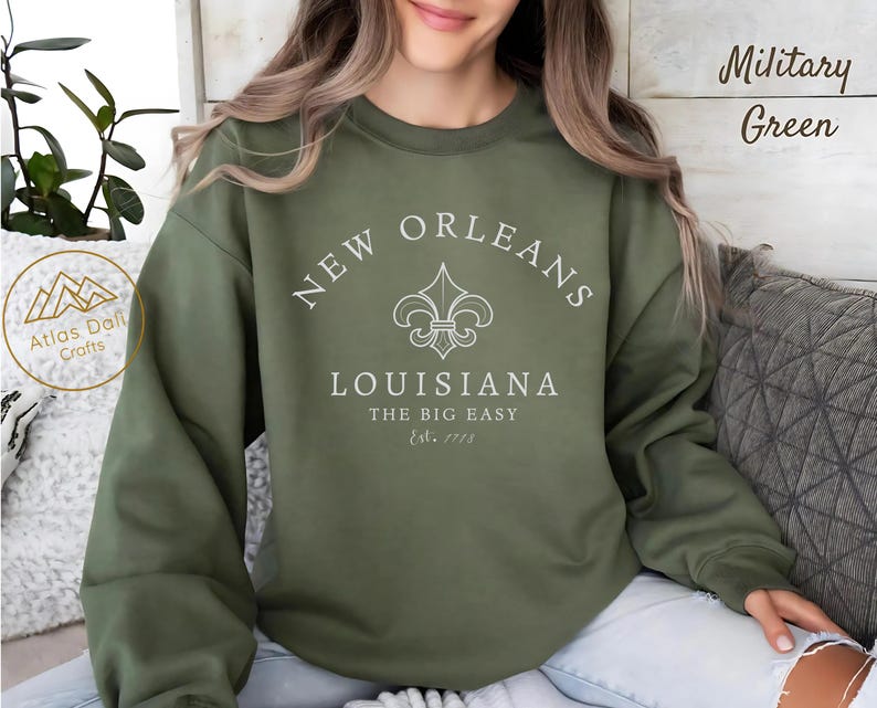 May include: Military green sweatshirt with the text "New Orleans Louisiana The Big Easy Est. 1718" in white. The sweatshirt features a fleur-de-lis design. The brand name "Atlas Daffi Crafts" is visible on the left side.