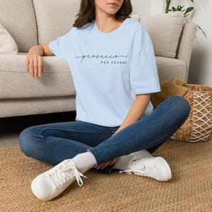 Op de afbeelding: Een lichtblauw T-shirt met korte mouwen en de zwarte tekst "prosecco PER FAVORE" op de borst gedrukt. Het shirt heeft een ronde hals en een relaxte pasvorm, gecombineerd met een donkerblauwe spijkerbroek en witte sneakers met witte veters.