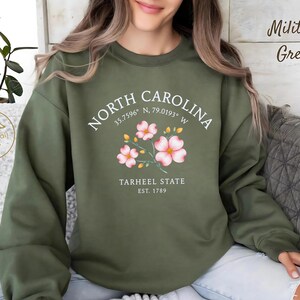 Op de afbeelding: Militairgroen sweatshirt met de tekst "NORTH CAROLINA" en co&ouml;rdinaten, plus "TARHEEL STATE EST. 1789". Het ontwerp bevat roze bloemen. Het sweatshirt is een casual stijl.