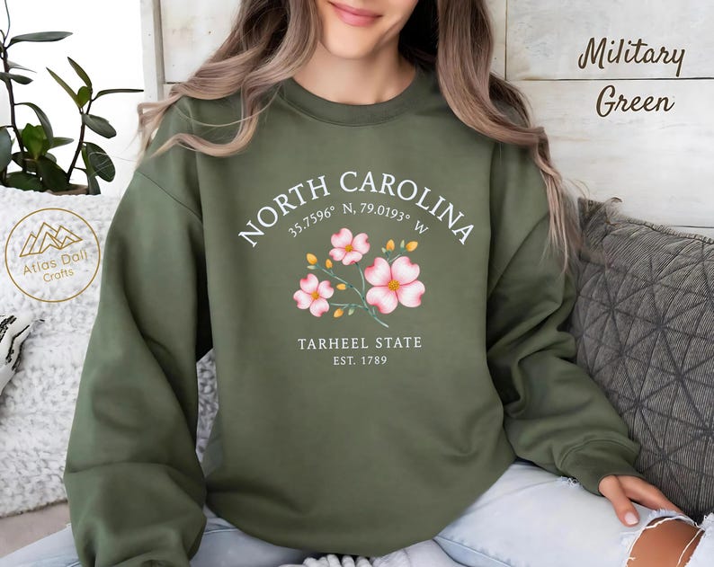 Op de afbeelding: Militairgroen sweatshirt met de tekst "NORTH CAROLINA" en co&ouml;rdinaten, plus "TARHEEL STATE EST. 1789". Het ontwerp bevat roze bloemen. Het sweatshirt is een casual stijl.