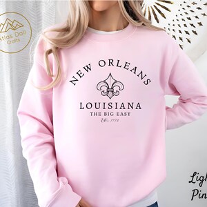 May include: Light pink sweatshirt with the text "NEW ORLEANS LOUISIANA THE BIG EASY Est. 1718" in black. The design includes a fleur-de-lis symbol. The sweatshirt is a light pink color.
