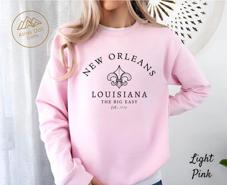 May include: Light pink sweatshirt with the text "NEW ORLEANS LOUISIANA THE BIG EASY Est. 1718" in black. The design includes a fleur-de-lis symbol. The sweatshirt is a light pink color.