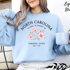 Op de afbeelding: Lichtblauw sweatshirt met de tekst "NORTH CAROLINA" en co&ouml;rdinaten, een bloemenafbeelding en "TARHEEL STATE EST. 1789". Het sweatshirt heeft ook de tekst "Light Blue" in de rechterbovenhoek.