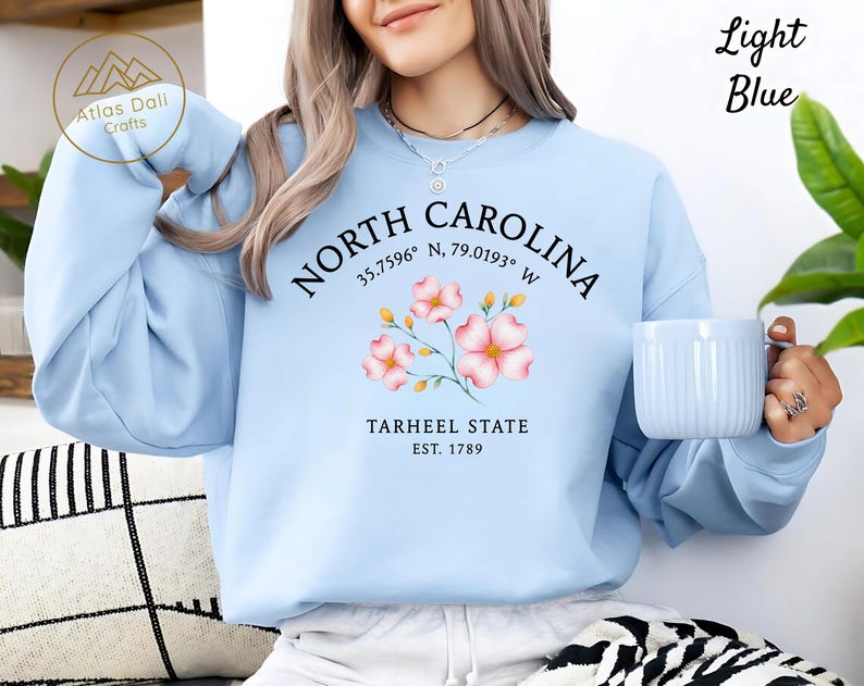 Op de afbeelding: Lichtblauw sweatshirt met de tekst "NORTH CAROLINA" en co&ouml;rdinaten, een bloemenafbeelding en "TARHEEL STATE EST. 1789". Het sweatshirt heeft ook de tekst "Light Blue" in de rechterbovenhoek.