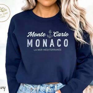 May include: Navy blue sweatshirt with the text "Monte Carlo MONACO" in white, along with an anchor graphic. The phrase "LA MER M&Eacute;DITERRAN&Eacute;E" is below the city name. The sweatshirt has a crew neck and long sleeves.
