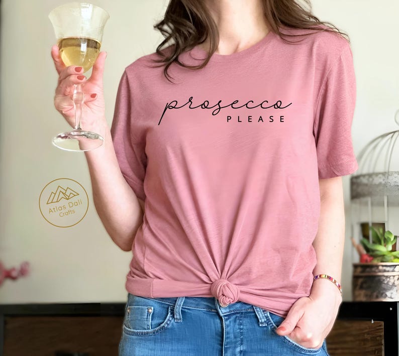 Peut inclure: Un t-shirt rose poudr&eacute; avec l'inscription "prosecco PLEASE" en &eacute;criture noire. Le t-shirt est nou&eacute; &agrave; la taille et associ&eacute; &agrave; un jean bleu. Une personne tient un verre de vin avec un liquide dor&eacute;.