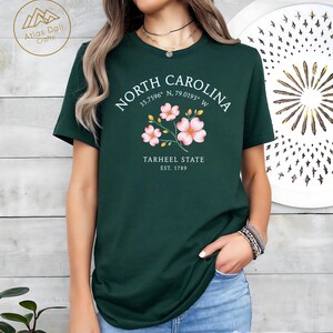 Może przedstawiać: Ciemnozielona koszulka z białym napisem "NORTH CAROLINA", wsp&oacute;łrzędnymi i "TARHEEL STATE EST. 1789". Koszulka ma r&oacute;wnież kwiatowy wz&oacute;r z r&oacute;żowymi kwiatami.