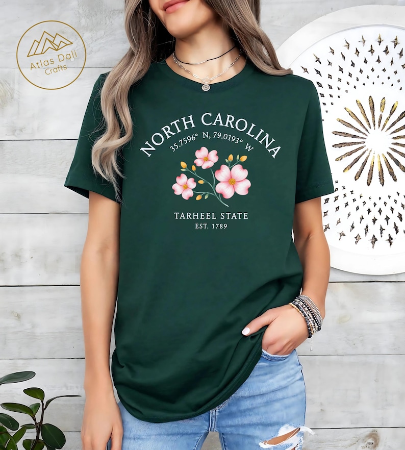 Może przedstawiać: Ciemnozielona koszulka z białym napisem "NORTH CAROLINA", wsp&oacute;łrzędnymi i "TARHEEL STATE EST. 1789". Koszulka ma r&oacute;wnież kwiatowy wz&oacute;r z r&oacute;żowymi kwiatami.