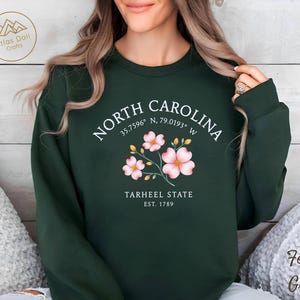 Op de afbeelding: Bostongroen sweatshirt met de tekst "NORTH CAROLINA" en co&ouml;rdinaten, plus een bloemenafbeelding. Daaronder staat "TARHEEL STATE EST. 1789". De sweatshirt is effen van kleur en heeft een ronde hals.