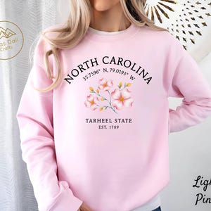 Op de afbeelding: Lichtroze sweatshirt met de tekst "NORTH CAROLINA" en co&ouml;rdinaten, een bloemenontwerp en "TARHEEL STATE EST. 1789". Het sweatshirt heeft ook de tekst "Light Pink" rechtsonder.
