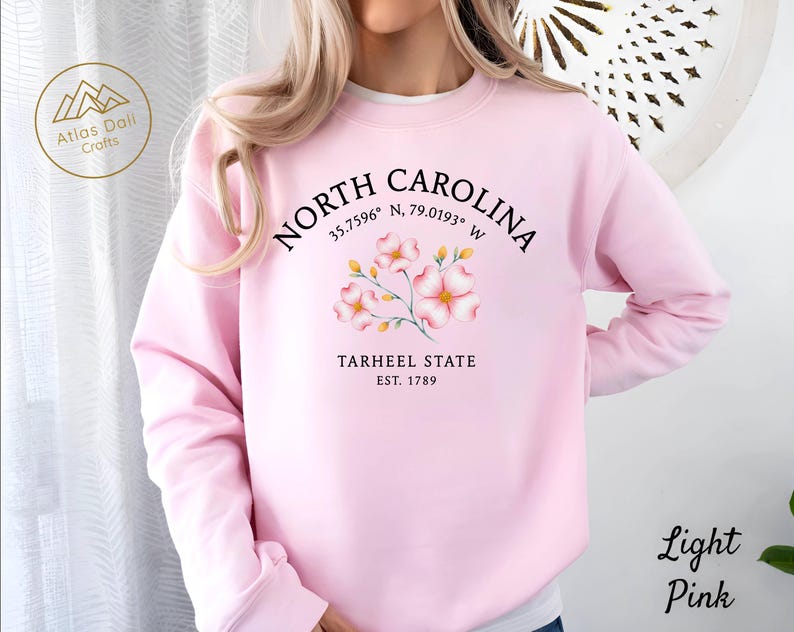 Op de afbeelding: Lichtroze sweatshirt met de tekst "NORTH CAROLINA" en co&ouml;rdinaten, een bloemenontwerp en "TARHEEL STATE EST. 1789". Het sweatshirt heeft ook de tekst "Light Pink" rechtsonder.