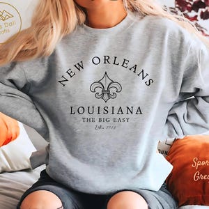 May include: A light grey sweatshirt with the words "NEW ORLEANS" arched across the top, a fleur-de-lis emblem, and "LOUISIANA" below. The text "THE BIG EASY" and "Est. 1718" are also printed on the sweatshirt. The sweatshirt is labeled "Sport Grey".