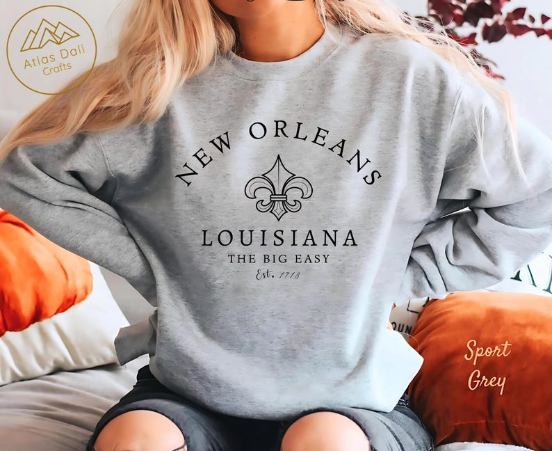May include: A light grey sweatshirt with the words "NEW ORLEANS" arched across the top, a fleur-de-lis emblem, and "LOUISIANA" below. The text "THE BIG EASY" and "Est. 1718" are also printed on the sweatshirt. The sweatshirt is labeled "Sport Grey".