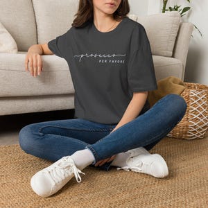 Op de afbeelding: Een donkergrijs T-shirt met korte mouwen en de witte tekst "prosecco PER FAVORE" op de borst. Het shirt heeft een ronde hals en een relaxte pasvorm, gecombineerd met een blauwe spijkerbroek en witte vetersneakers met witte sokken.