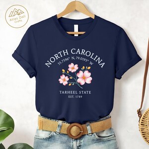 Może przedstawiać: Granatowa koszulka z kwiatowym wzorem i napisem "NORTH CAROLINA" nad "TARHEEL STATE EST. 1789". Koszulka ma podwinięte rękawy i jest połączona z brązowym paskiem i niebieskimi dżinsami.