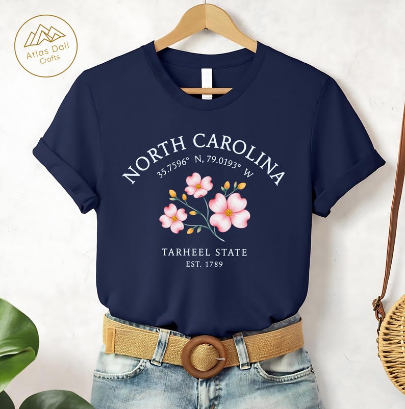 Może przedstawiać: Granatowa koszulka z kwiatowym wzorem i napisem "NORTH CAROLINA" nad "TARHEEL STATE EST. 1789". Koszulka ma podwinięte rękawy i jest połączona z brązowym paskiem i niebieskimi dżinsami.