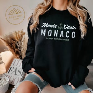May include: Black sweatshirt with the text "Monte Carlo MONACO LA MER M&Eacute;DITERRAN&Eacute;E" in white. An anchor graphic is between "Monte Carlo" and "MONACO". The sweatshirt has long sleeves and a crew neck. The brand logo is in the upper left corner.