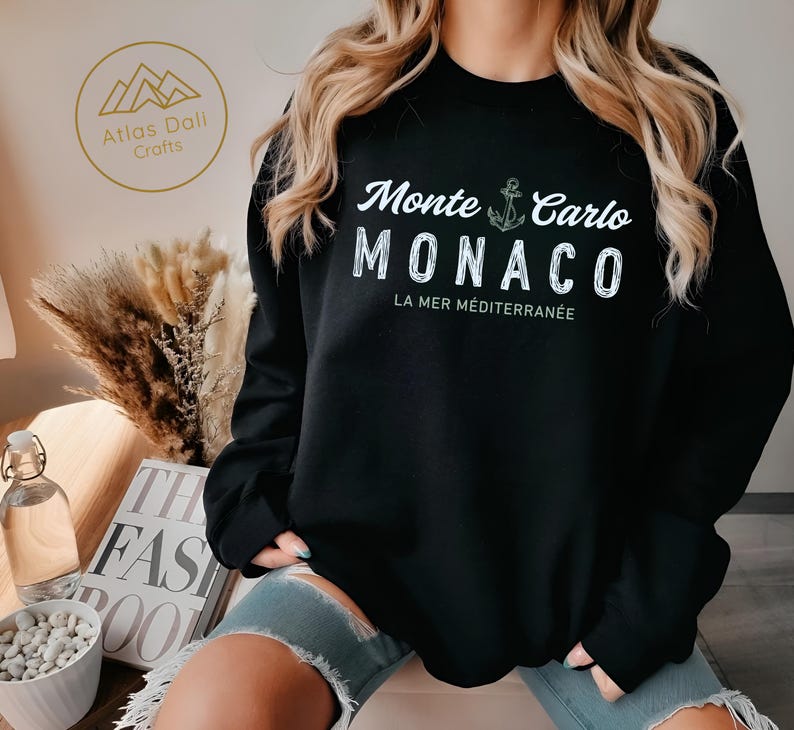 May include: Black sweatshirt with the text "Monte Carlo MONACO LA MER M&Eacute;DITERRAN&Eacute;E" in white. An anchor graphic is between "Monte Carlo" and "MONACO". The sweatshirt has long sleeves and a crew neck. The brand logo is in the upper left corner.
