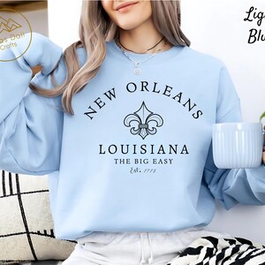 May include: Light blue sweatshirt with "New Orleans Louisiana" text and a fleur-de-lis graphic. The text "The Big Easy" and "Est. 1718" are also printed on the sweatshirt. A light blue mug is held in the hand.