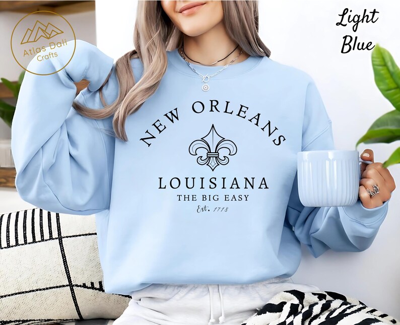 May include: Light blue sweatshirt with "New Orleans Louisiana" text and a fleur-de-lis graphic. The text "The Big Easy" and "Est. 1718" are also printed on the sweatshirt. A light blue mug is held in the hand.