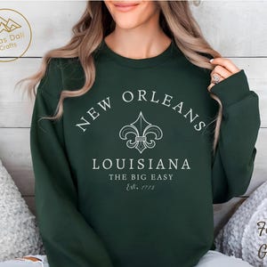 May include: Forest green sweatshirt with the text "NEW ORLEANS LOUISIANA THE BIG EASY Est. 1718" in white. The design includes a fleur-de-lis symbol. The sweatshirt is a crew neck style.