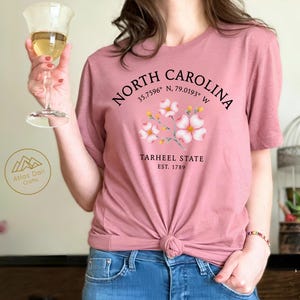Może przedstawiać: Pyłowa r&oacute;żowa koszulka z napisem "NORTH CAROLINA", wsp&oacute;łrzędnymi i "TARHEEL STATE EST. 1789". Koszulka ma kwiatowy wz&oacute;r i jest zawiązana w talii. Osoba trzyma kieliszek wina.