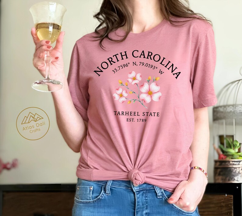 Może przedstawiać: Pyłowa r&oacute;żowa koszulka z napisem "NORTH CAROLINA", wsp&oacute;łrzędnymi i "TARHEEL STATE EST. 1789". Koszulka ma kwiatowy wz&oacute;r i jest zawiązana w talii. Osoba trzyma kieliszek wina.