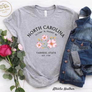 Może przedstawiać: Szara koszulka typu athletic heather z napisem "NORTH CAROLINA", kwiatowym wzorem i "TARHEEL STATE EST. 1789". Kurtka dżinsowa jest zarzucona na koszulkę. Bukiet r&oacute;żowych r&oacute;ż i zieleni znajduje się po lewej stronie.