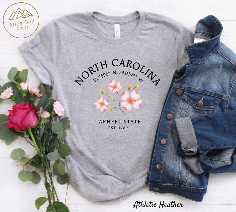 Może przedstawiać: Szara koszulka typu athletic heather z napisem "NORTH CAROLINA", kwiatowym wzorem i "TARHEEL STATE EST. 1789". Kurtka dżinsowa jest zarzucona na koszulkę. Bukiet r&oacute;żowych r&oacute;ż i zieleni znajduje się po lewej stronie.
