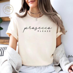 Peut inclure: T-shirt cr&egrave;me avec l'inscription "prosecco PLEASE" en &eacute;criture noire. Le t-shirt a des manches courtes et une coupe d&eacute;contract&eacute;e. Le mot "Natural" est imprim&eacute; en bas &agrave; droite de l'image.