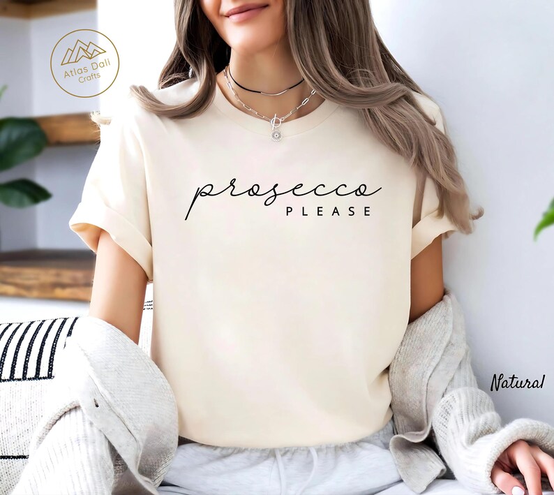 Peut inclure: T-shirt cr&egrave;me avec l'inscription "prosecco PLEASE" en &eacute;criture noire. Le t-shirt a des manches courtes et une coupe d&eacute;contract&eacute;e. Le mot "Natural" est imprim&eacute; en bas &agrave; droite de l'image.