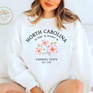 Op de afbeelding: Witte sweatshirt met de tekst "NORTH CAROLINA" en co&ouml;rdinaten, versierd met roze bloemmotieven. Daaronder staat "TARHEEL STATE EST. 1789". Een merklogo staat linksboven. Het woord "White" staat rechtsonder.