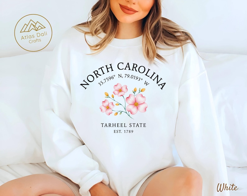 Op de afbeelding: Witte sweatshirt met de tekst "NORTH CAROLINA" en co&ouml;rdinaten, versierd met roze bloemmotieven. Daaronder staat "TARHEEL STATE EST. 1789". Een merklogo staat linksboven. Het woord "White" staat rechtsonder.