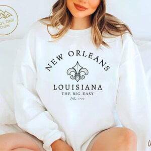 May include: White sweatshirt with the text "NEW ORLEANS LOUISIANA THE BIG EASY Est. 1718" in black. A fleur-de-lis symbol is centered above the text. The sweatshirt is a crew neck style.