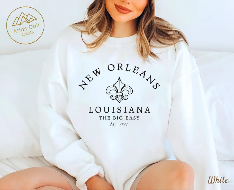 May include: White sweatshirt with the text "NEW ORLEANS LOUISIANA THE BIG EASY Est. 1718" in black. A fleur-de-lis symbol is centered above the text. The sweatshirt is a crew neck style.