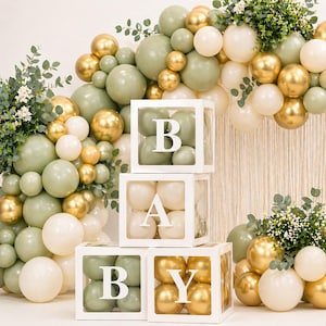 May include: A baby shower decoration with white boxes spelling "BABY" filled with balloons. The balloons are gold, white, and green, with greenery. The display is designed for a celebratory event.